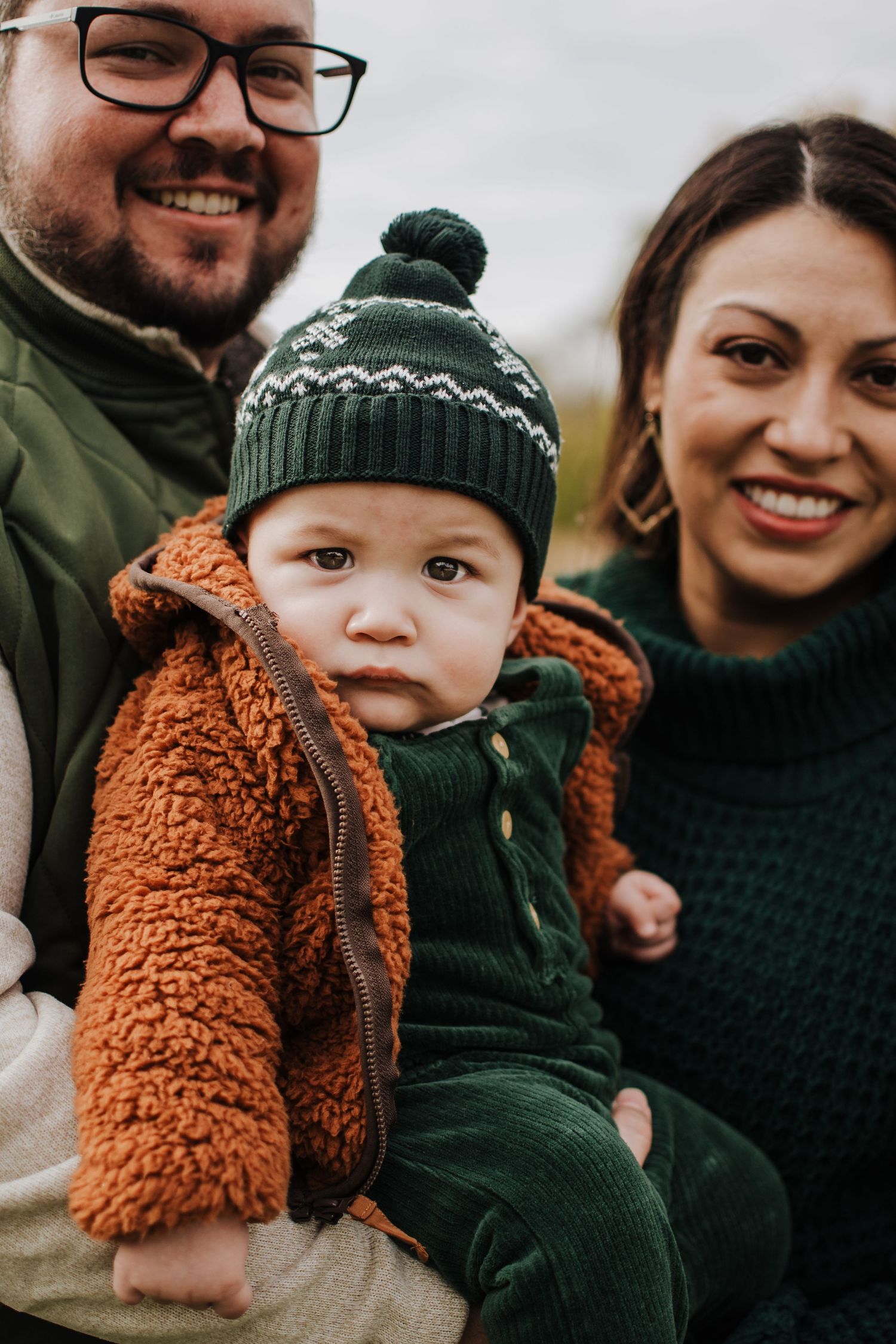 A cozy fall family photo featuring a baby in a brown sherpa jacket and green knit hat with parents outdoors.