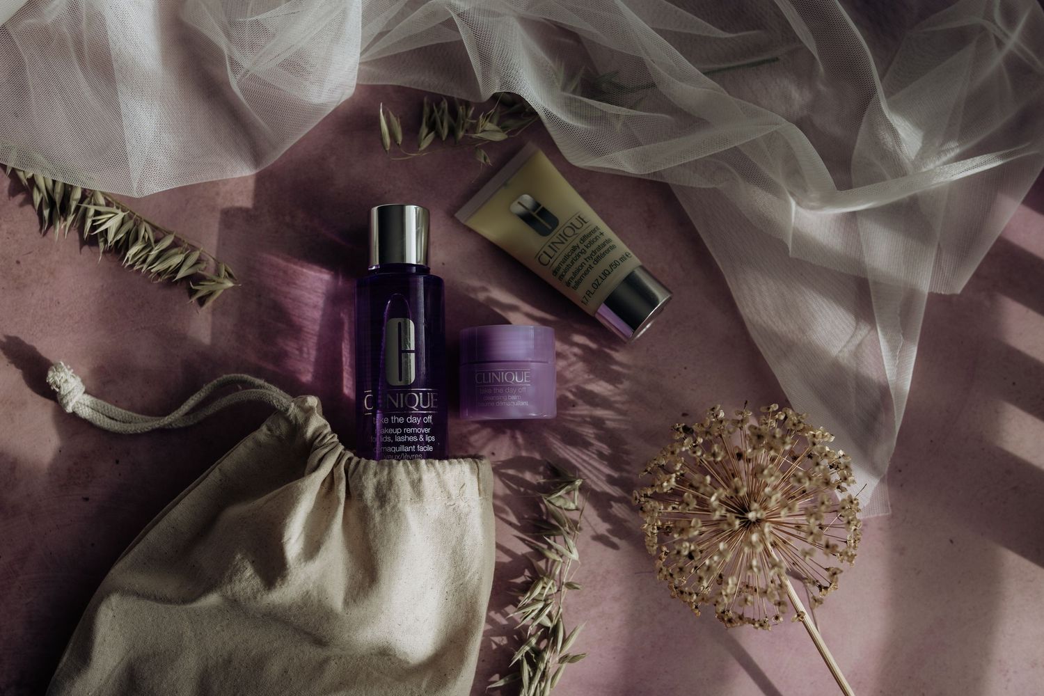 Beauty products and dried floral arrangement styled on pink silk fabric with soft white tulle draping.