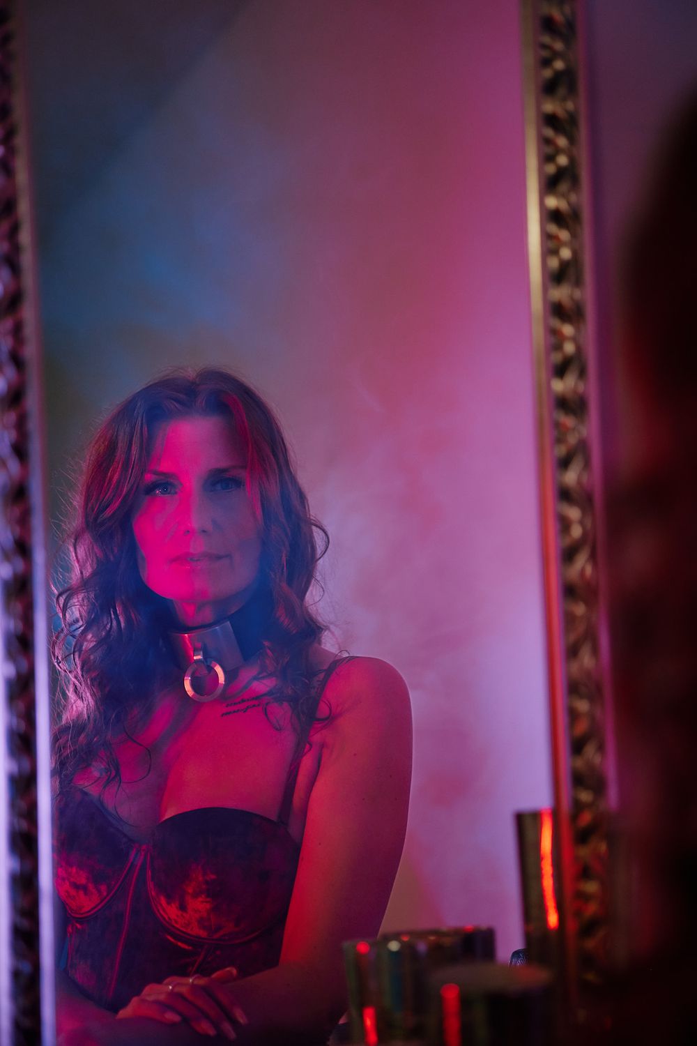 A person in dark attire photographed in moody red and blue lighting with an ornate frame visible in background.