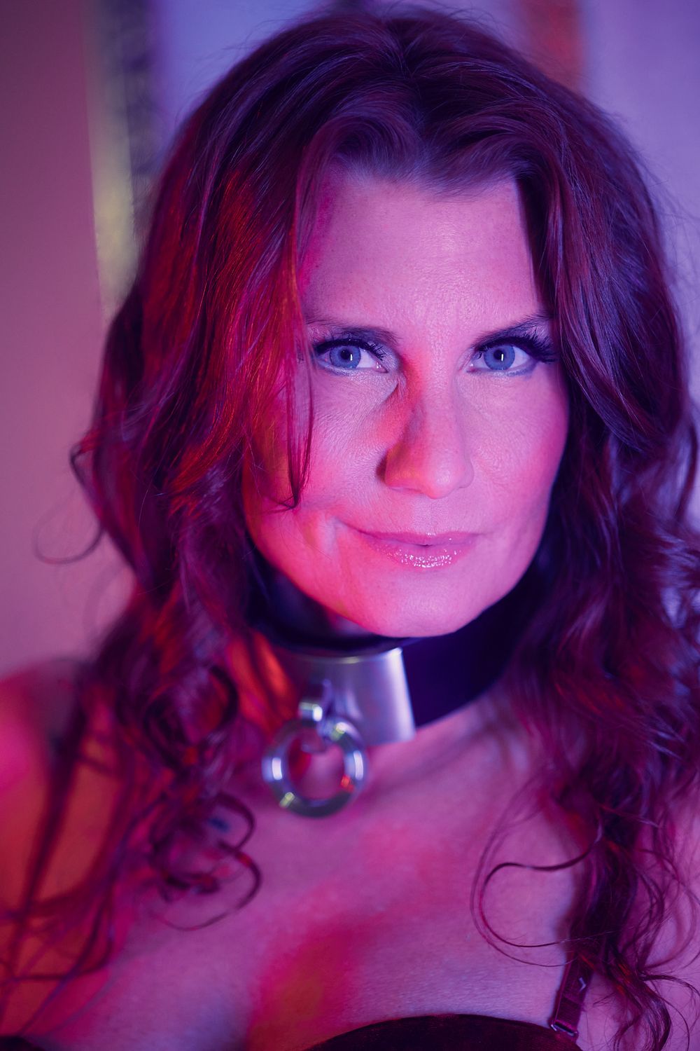 A portrait in purple and red lighting showing a person with long dark wavy hair smiling at the camera.
