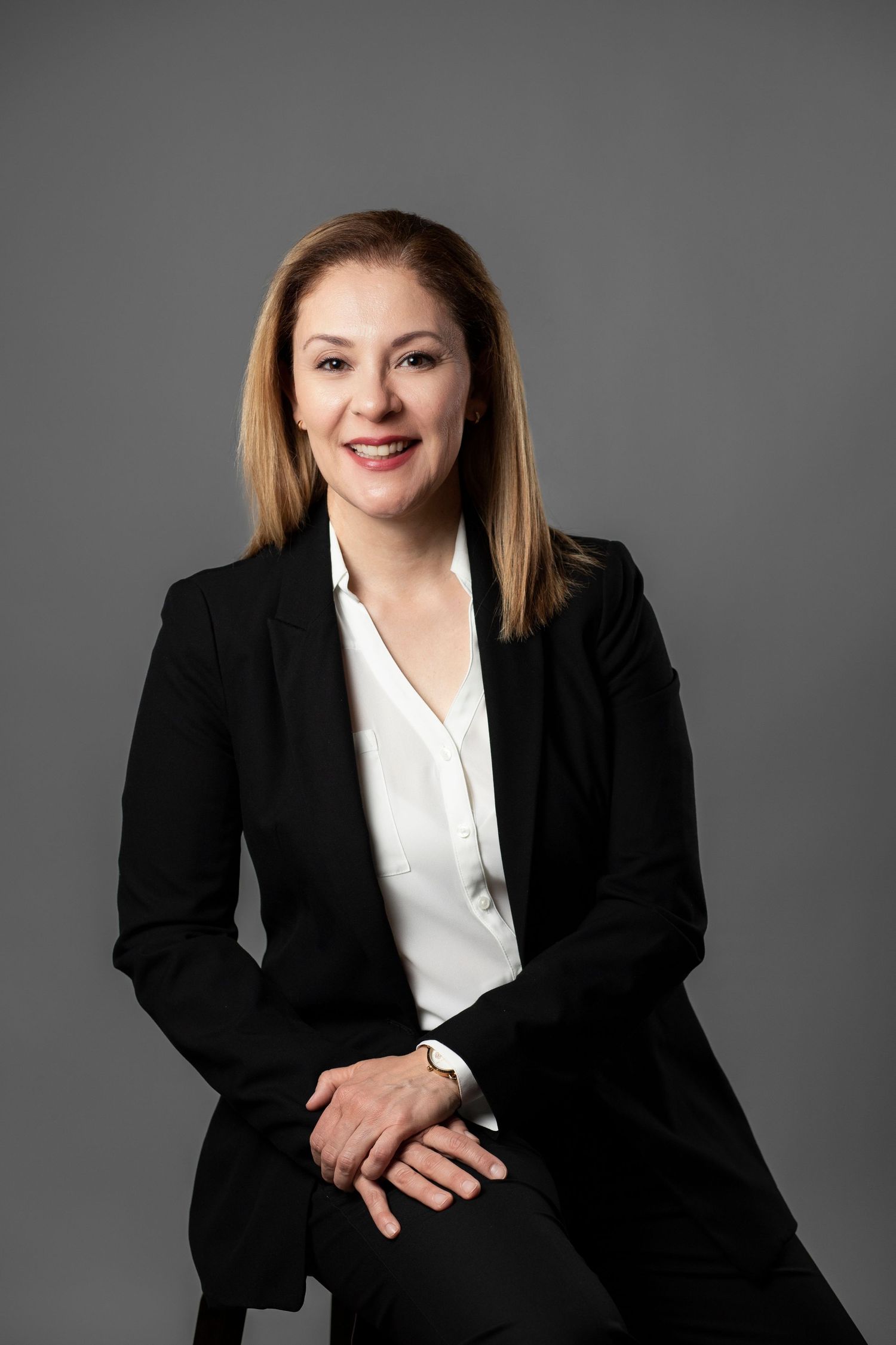 Business portrait of person in black suit and white blouse seated professionally against dark background.