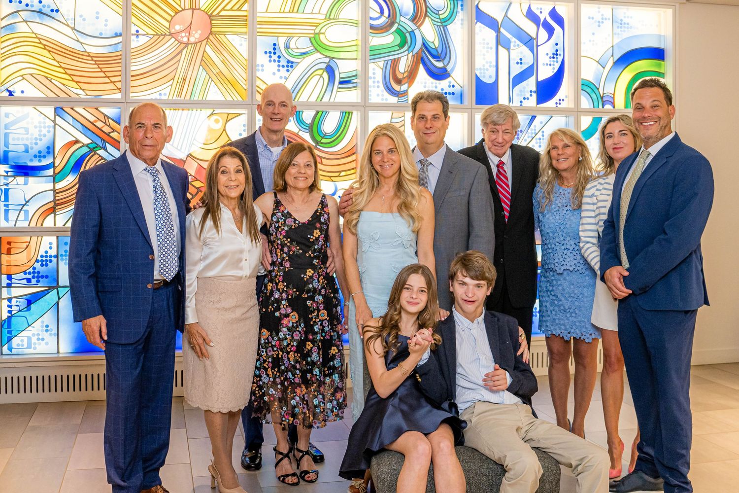 A group of people standing together in formal attire with colorful stained glass windows in the background.