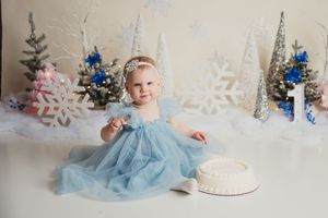 Winter cake smash photo session with light blue tulle dress, silver snowflakes, and sparkly Christmas trees in background.