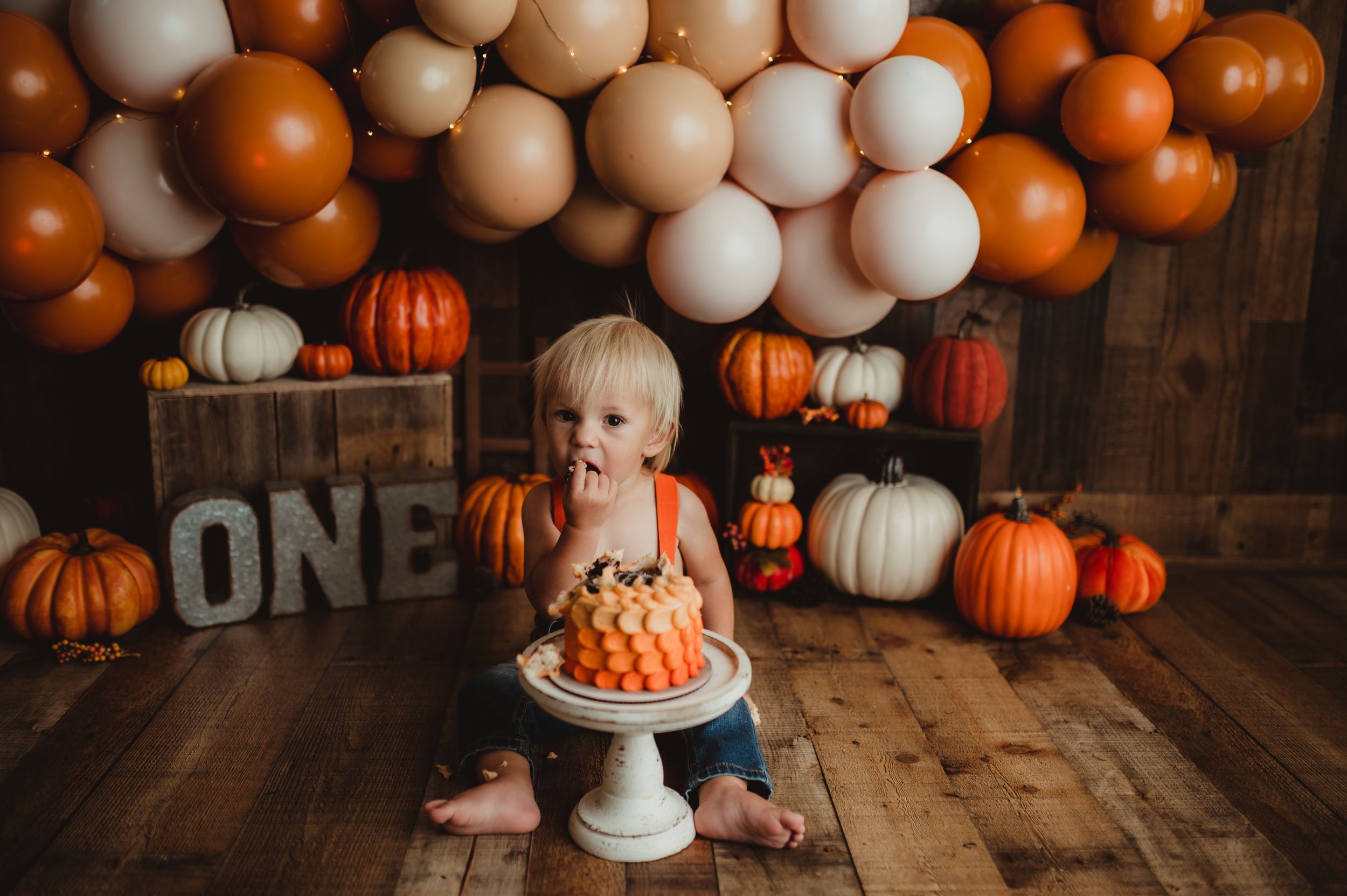 A baby in pink sits on a rustic wooden floor surrounded by orange and white pumpkins and fall decorations.