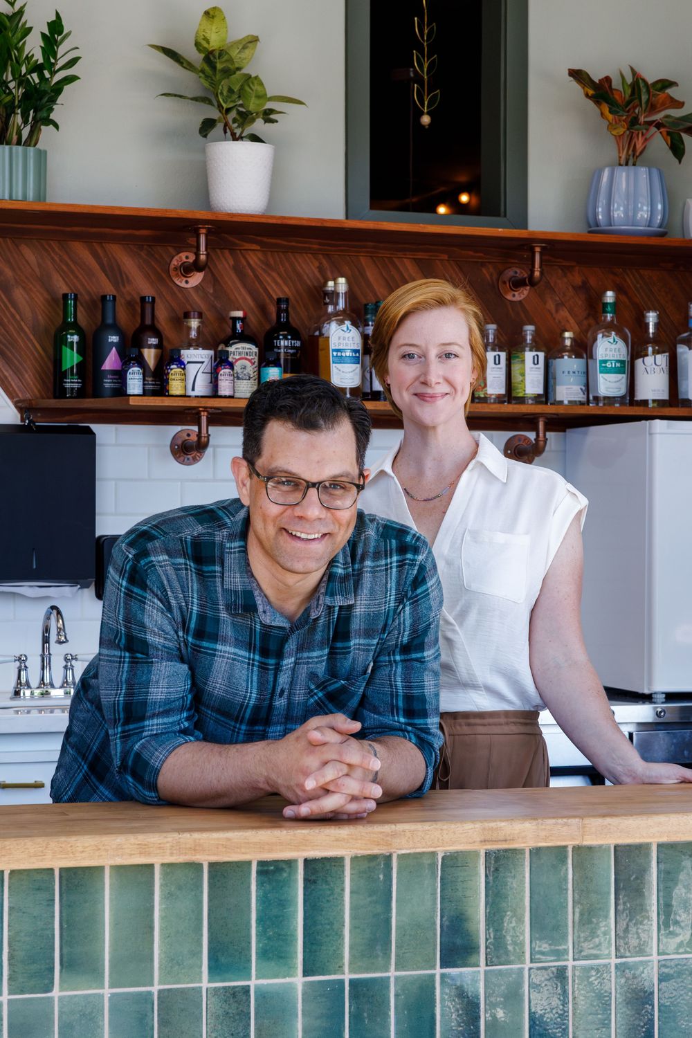 Two people sit at a tiled bar counter top in a kitchen setting with white walls and decorative houseplants.