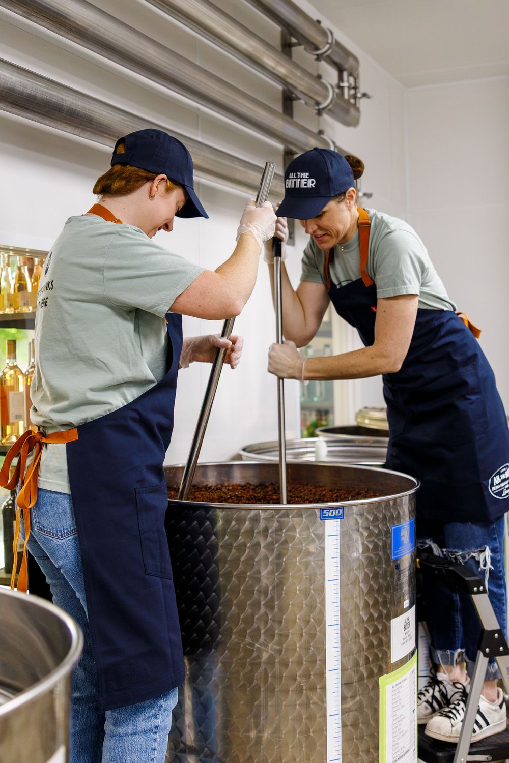 Two brewery workers stir a large batch of beer in stainless steel fermentation tanks at a craft brewery.
