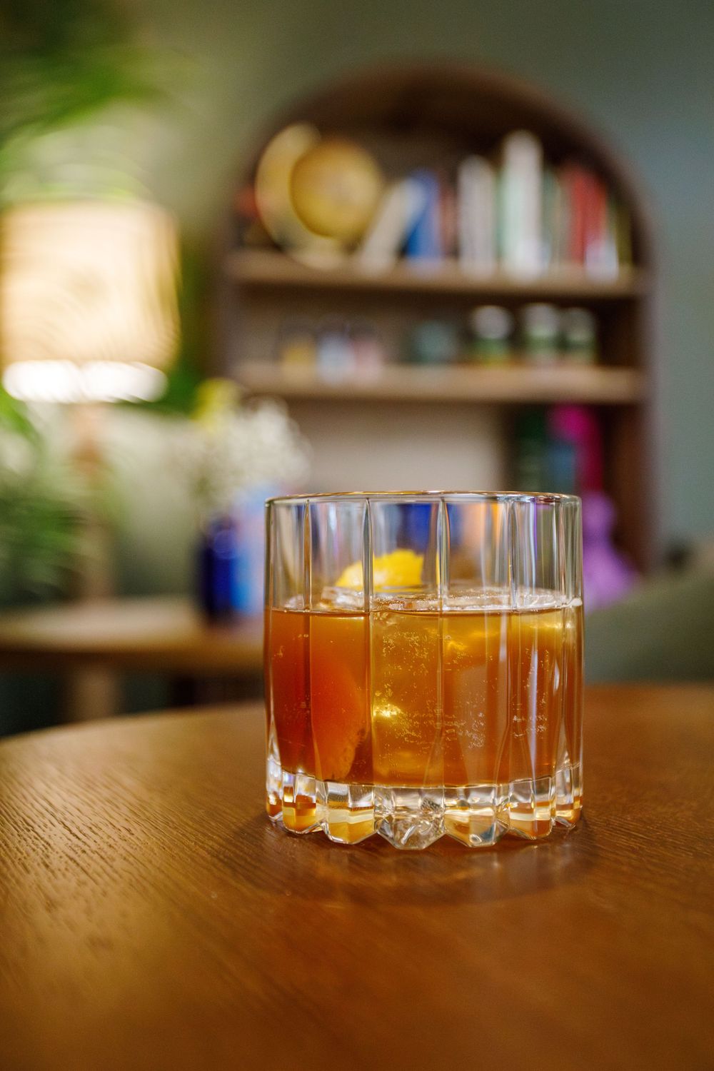 A golden-brown cocktail with ice in a crystal rocks glass sits on a wooden table with bookshelves visible in background.