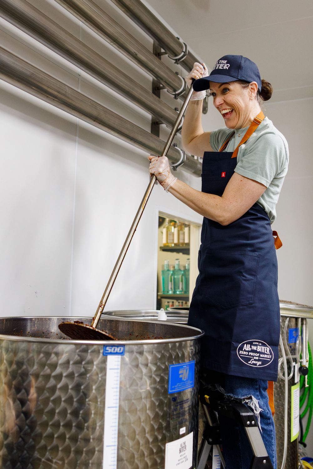 Worker enthusiastically stirring contents of a large steel fermentation tank at a craft brewery.