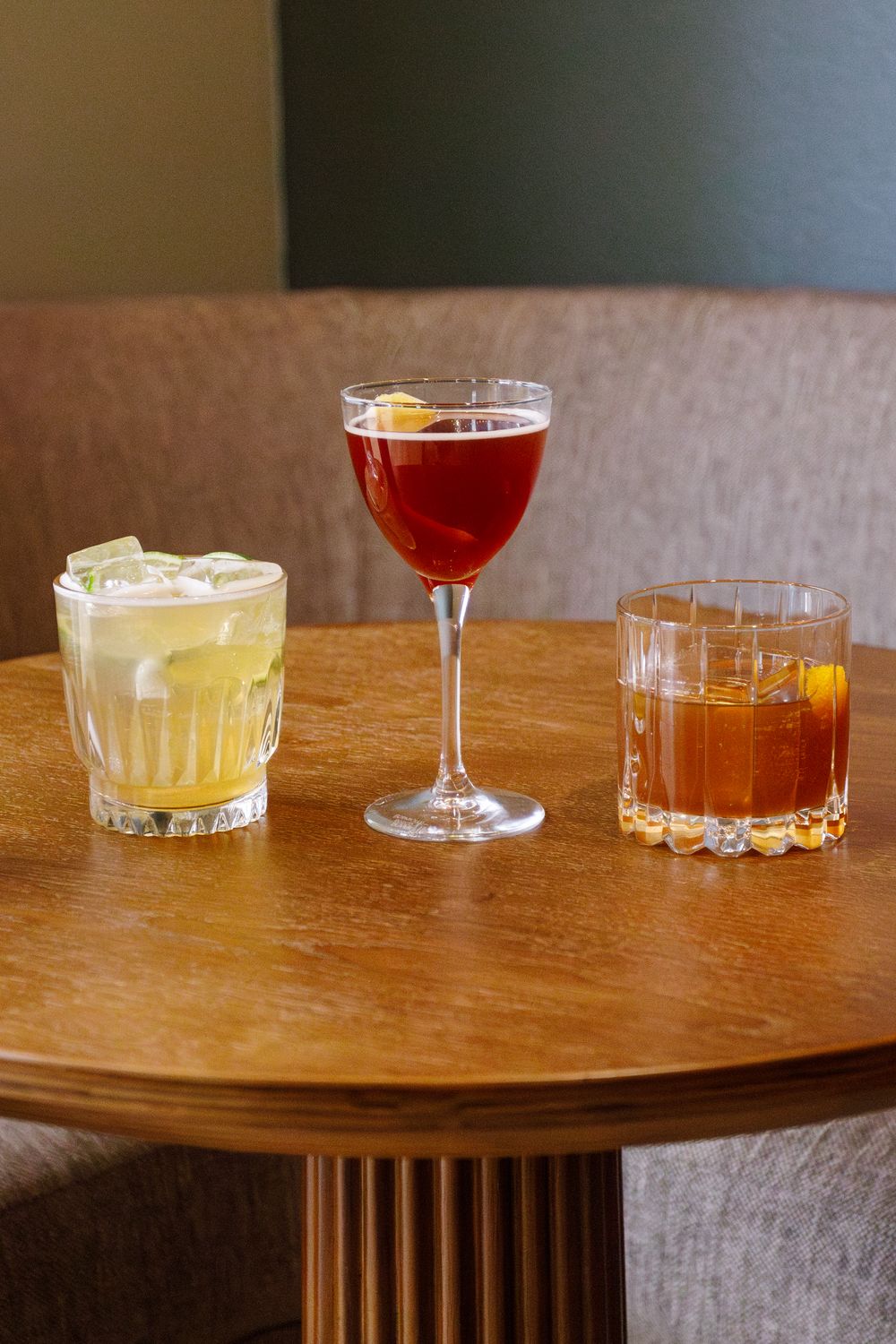 Three craft cocktails in different glasses arranged on a round wooden table.