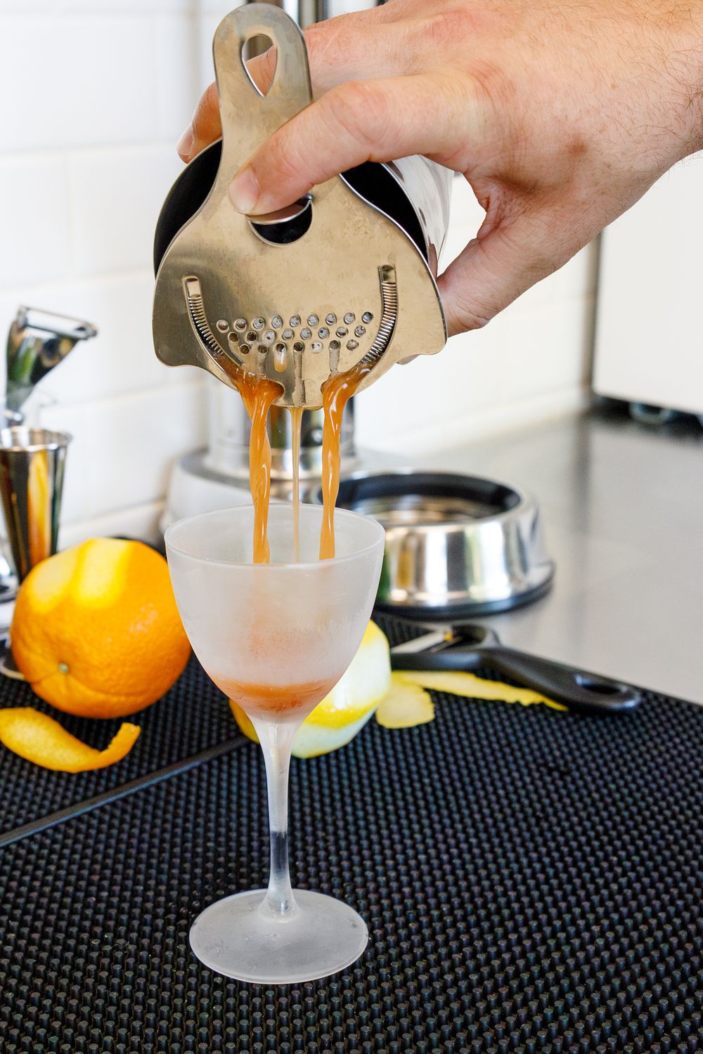 Bartender straining a cocktail into a martini glass with citrus fruits on a black bar mat.