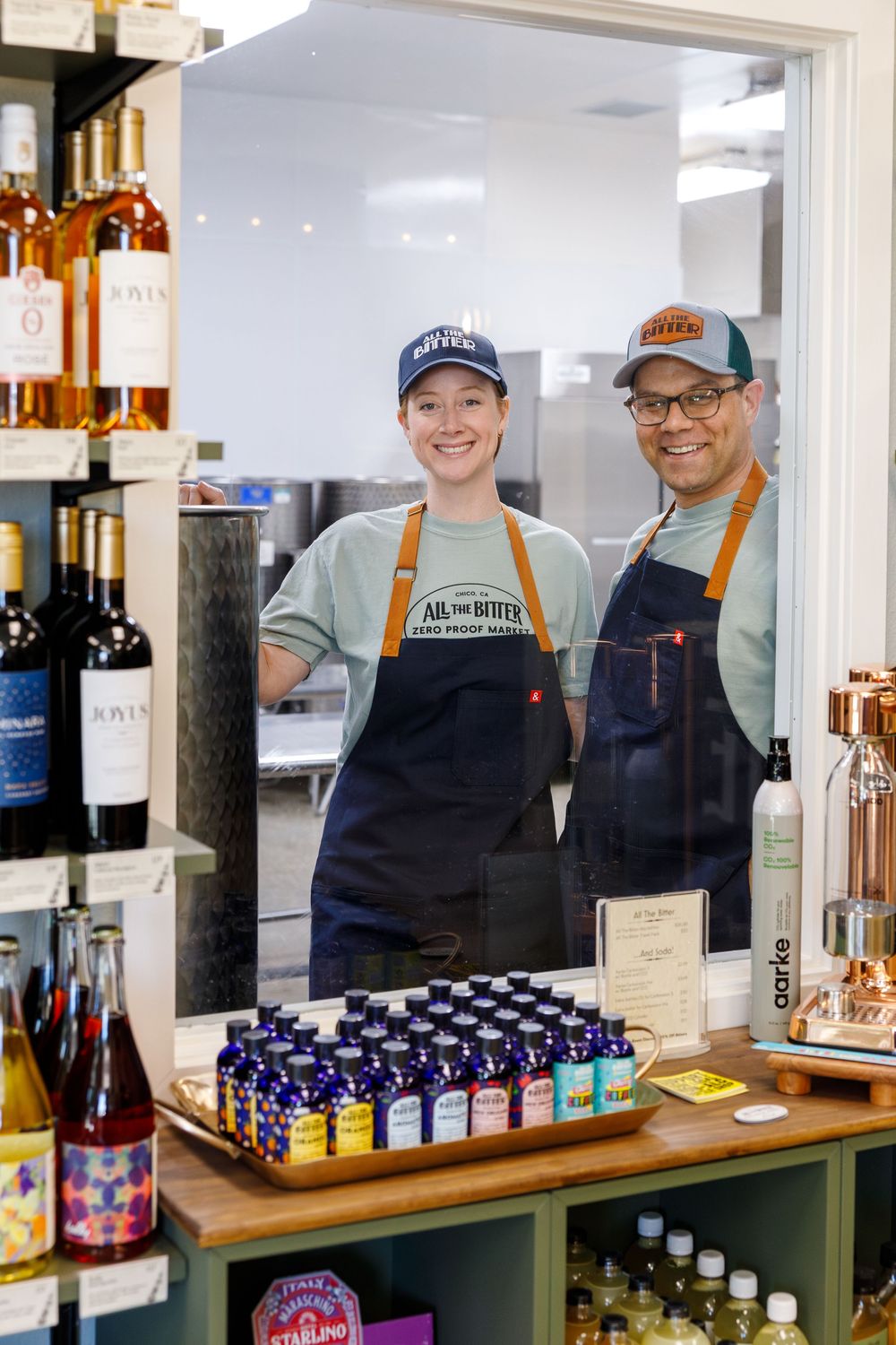 Two staff members in aprons stand behind a counter displaying bottles of spirits and cocktail ingredients at a bar.