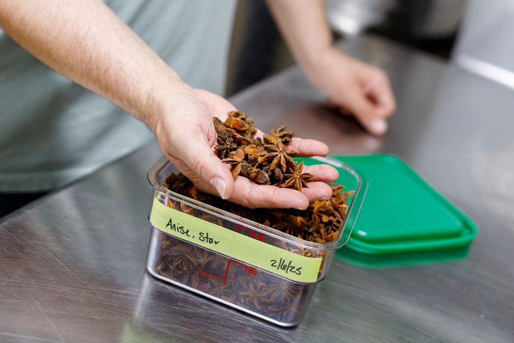 A hand holds dried star anise spices over an open glass storage jar with a green lid nearby.