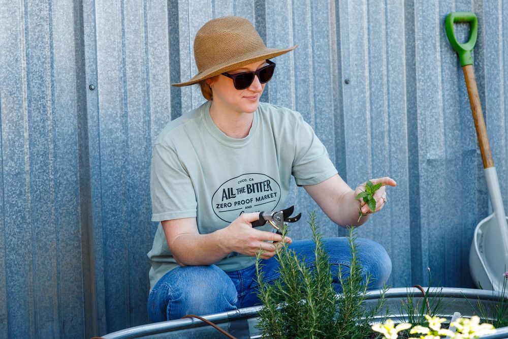 A gardener in a tan hat and gray shirt tends to plants while kneeling beside a metal wall.