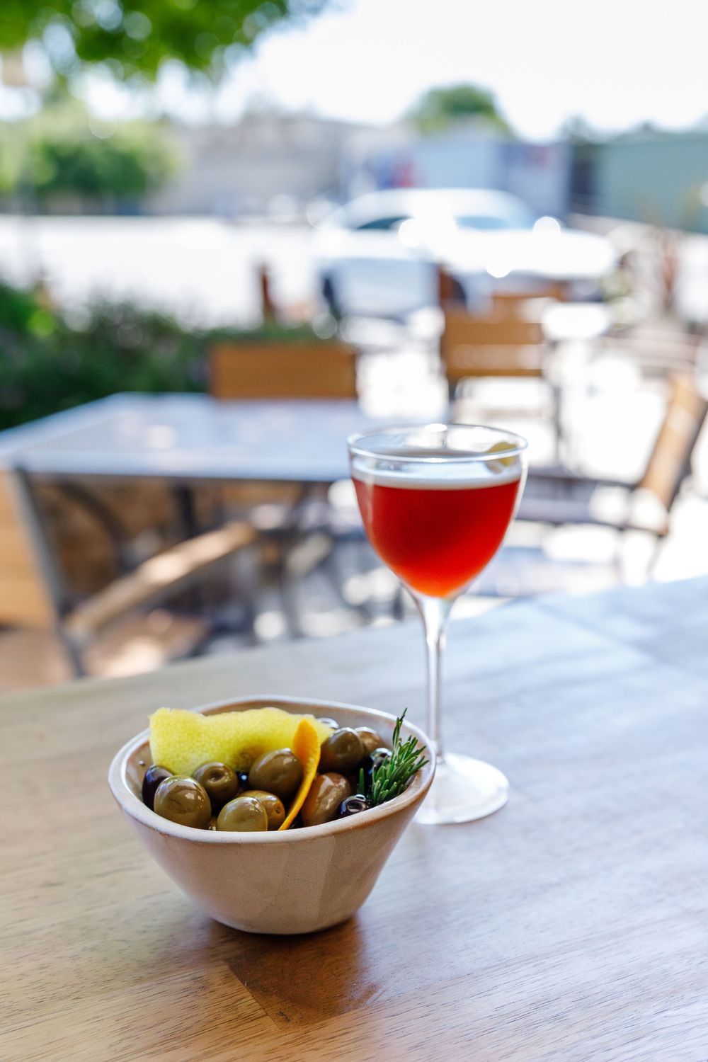 A glass of red wine sits beside a bowl of mixed olives and garnishes on an outdoor patio table.