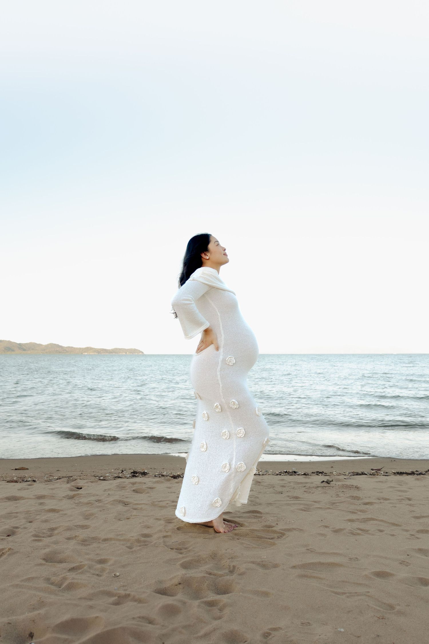 A person in a white dress poses elegantly on a sandy beach at sunset.