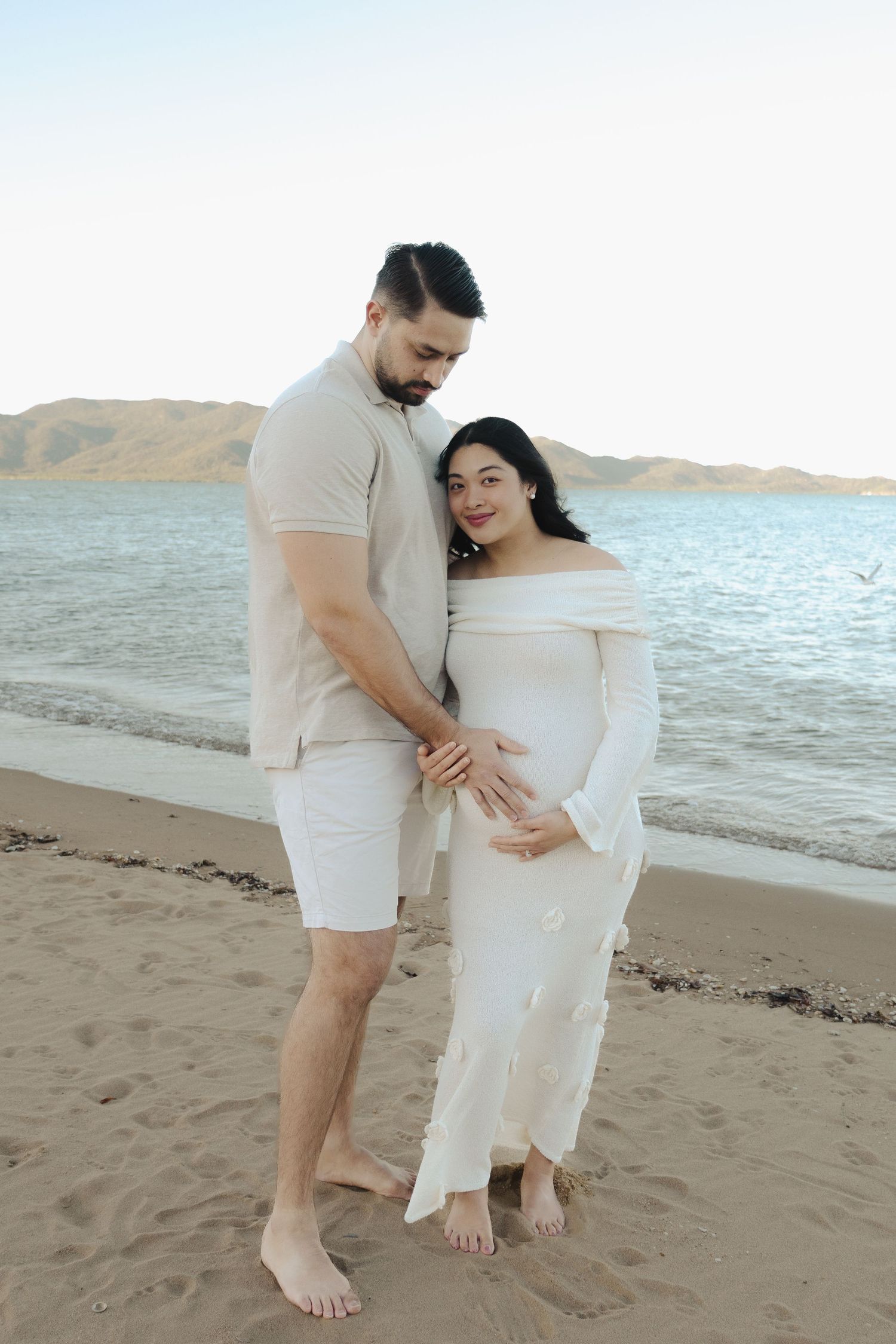 A couple shares a tender moment on the beach in coordinated white outfits.