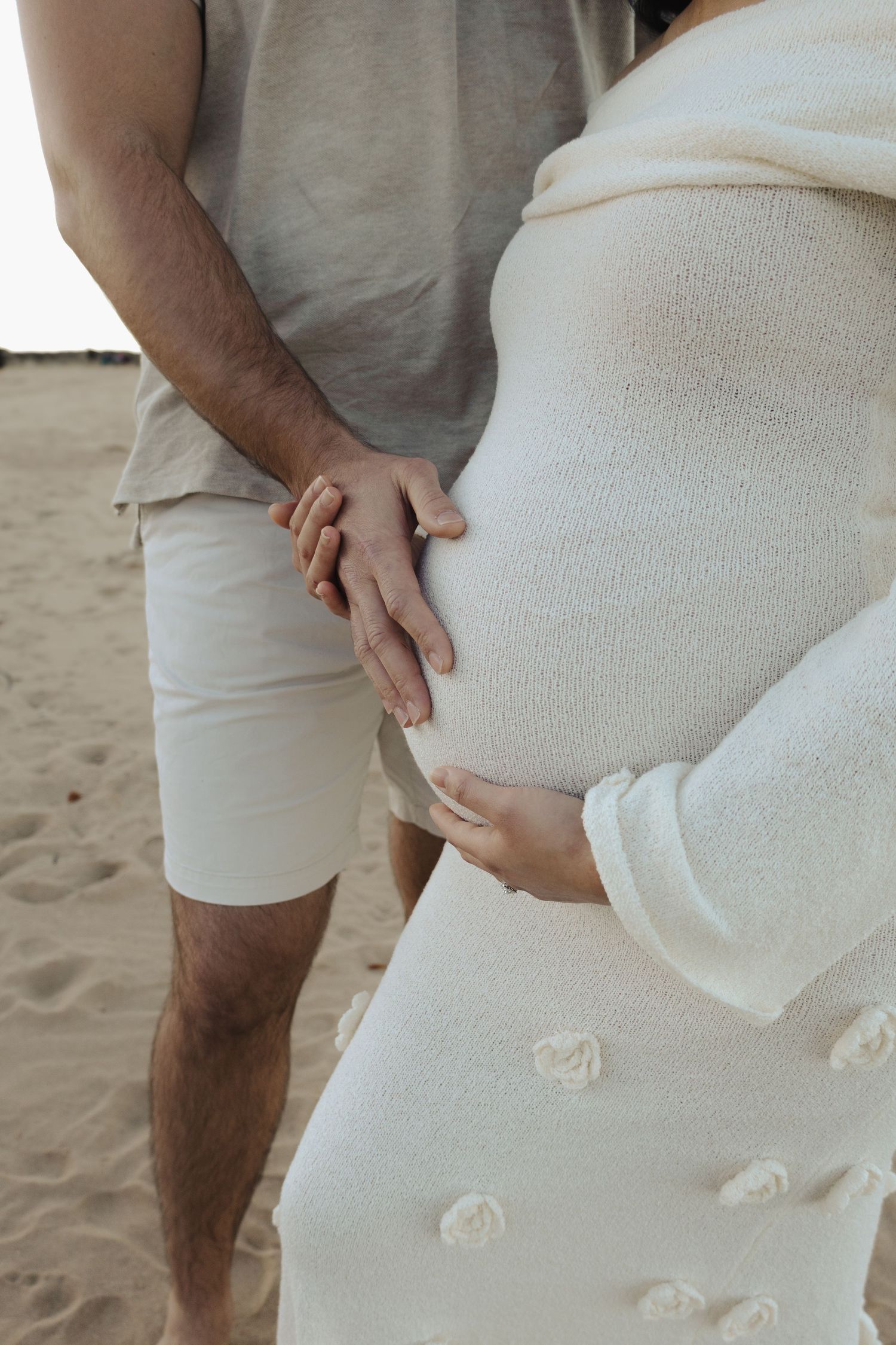 Close-up of a white maternity dress with delicate floral details against a sandy beach background.