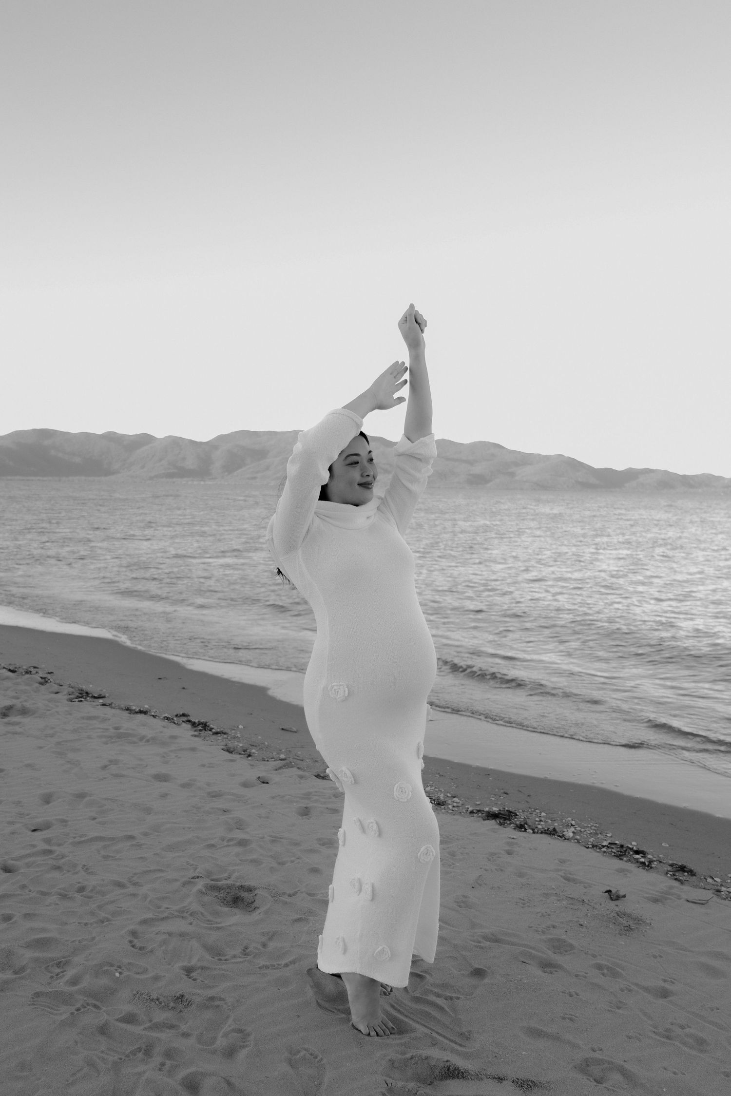Person in white dress dancing on the beach at sunset with ocean and mountains in background.