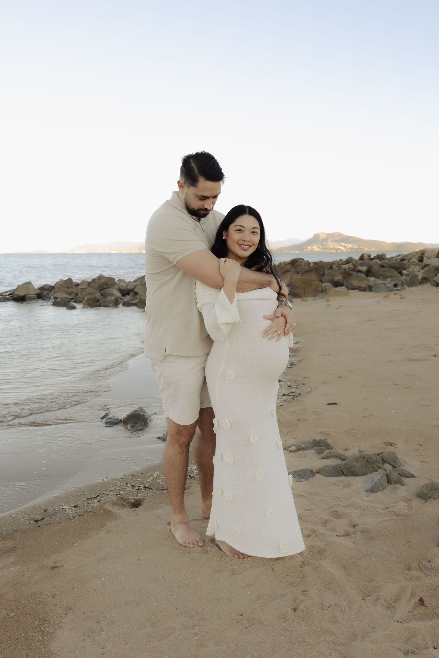 Romantic beach photoshoot with couple in white and beige outfits near rocky coastline.