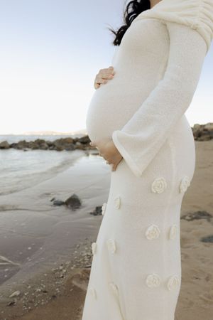 Side view of white maternity dress with floral appliques against ocean backdrop.