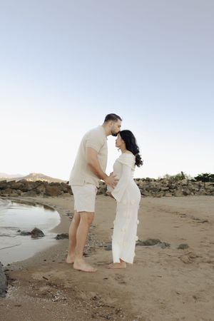 Couple sharing tender moment on sandy beach with rocky coastline in background.
