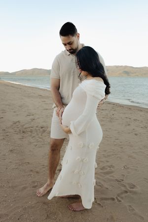 An intimate maternity photo showing a couple in light colored clothing on a sandy beach.
