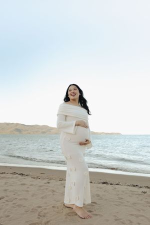 An expectant mother in a white dress smiles joyfully on a beach.