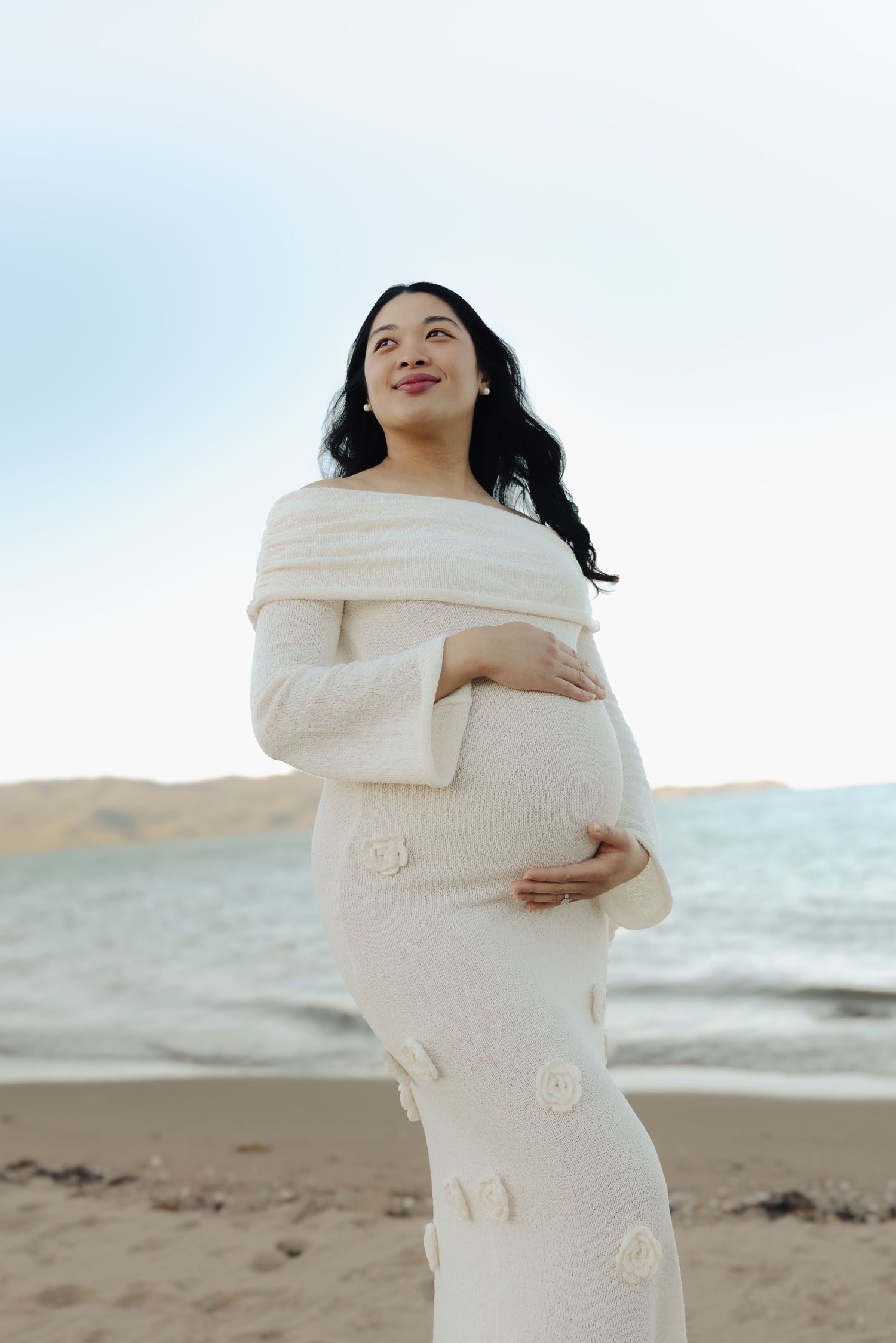 A radiant mother-to-be poses in an off-shoulder white dress by the ocean.