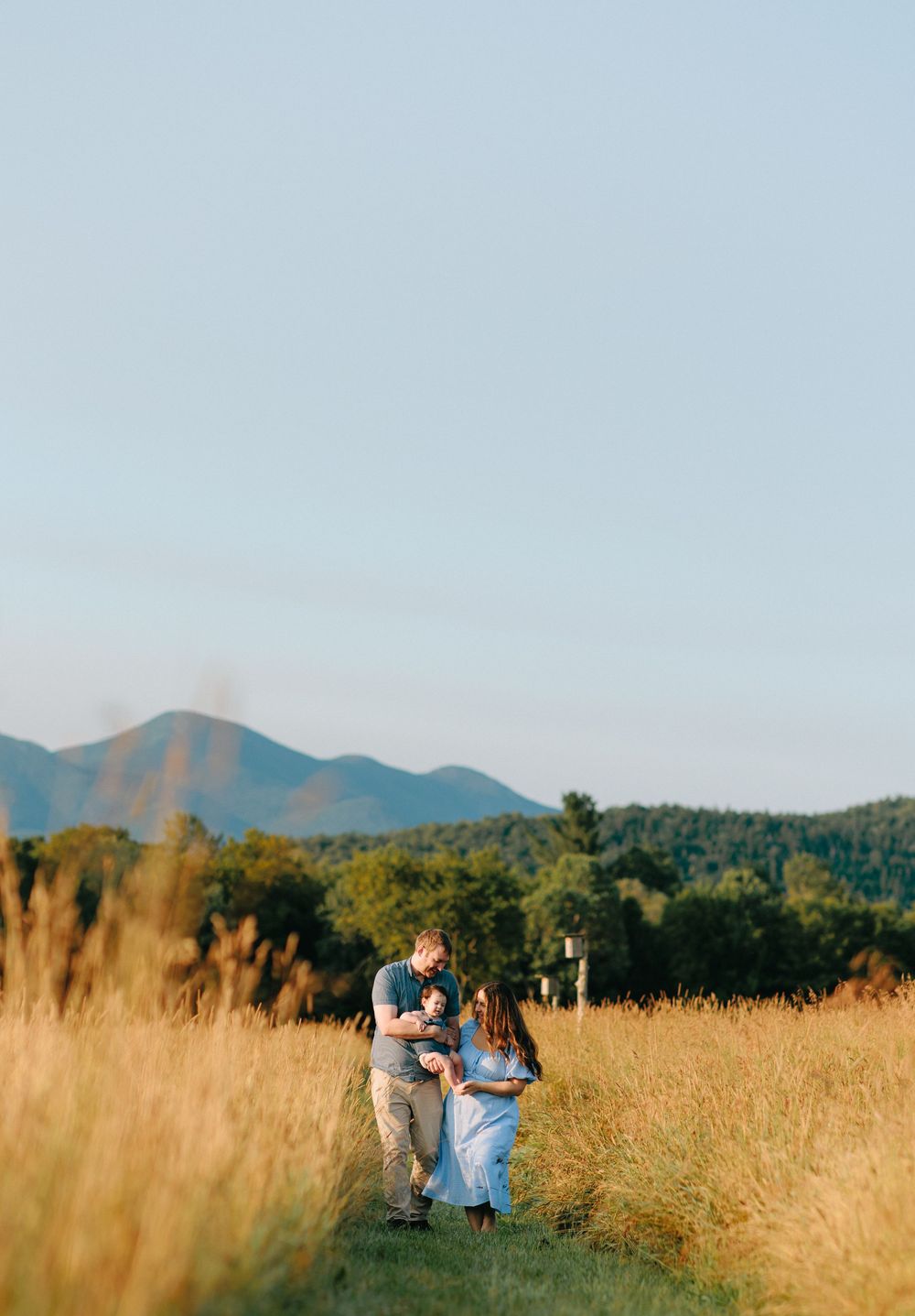 Two people share an intimate moment in a golden wheat field with mountain vistas and blue sky in the background.