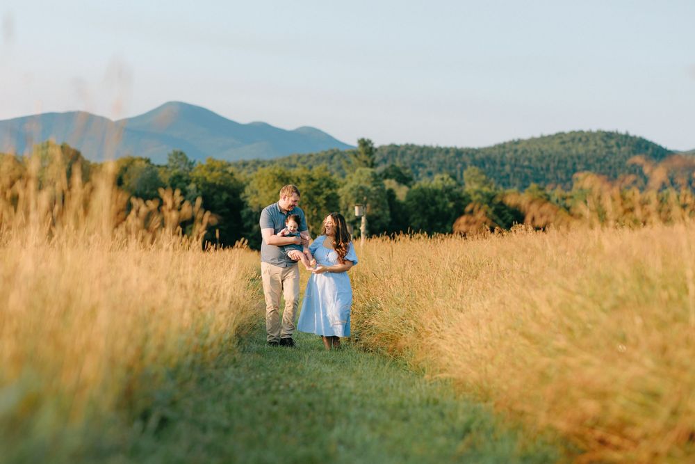 A couple walks along a path through golden wheatfields with mountains and forest in the background.
