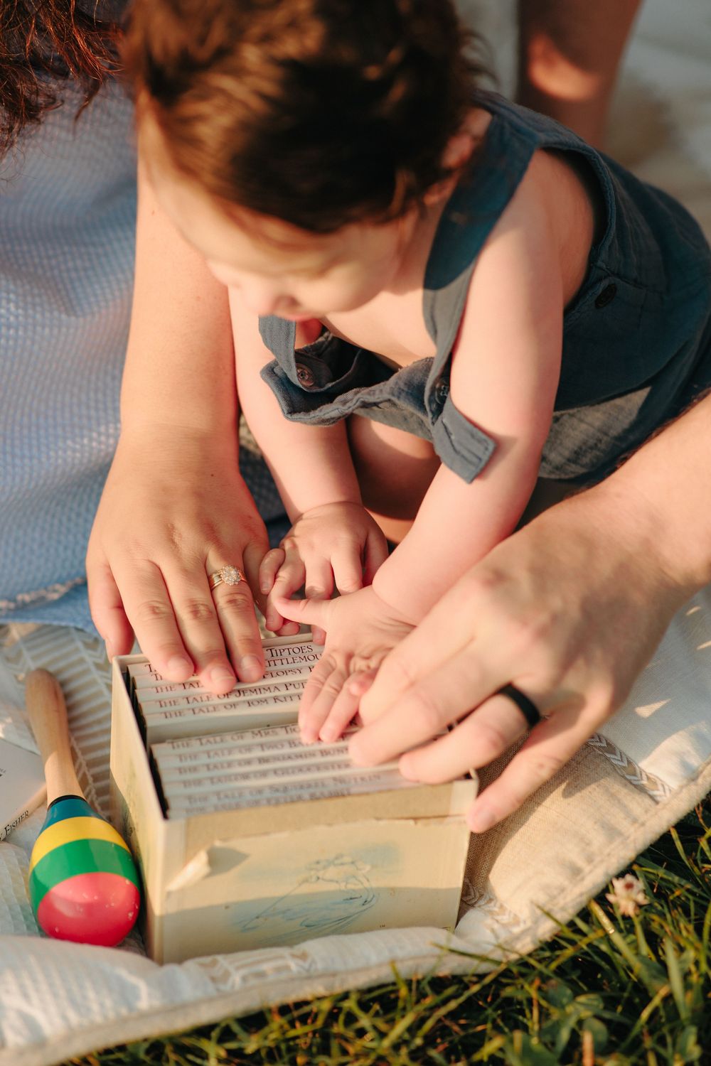 Reading a book together outdoors on a blanket with colorful toy blocks nearby during a sunny afternoon.