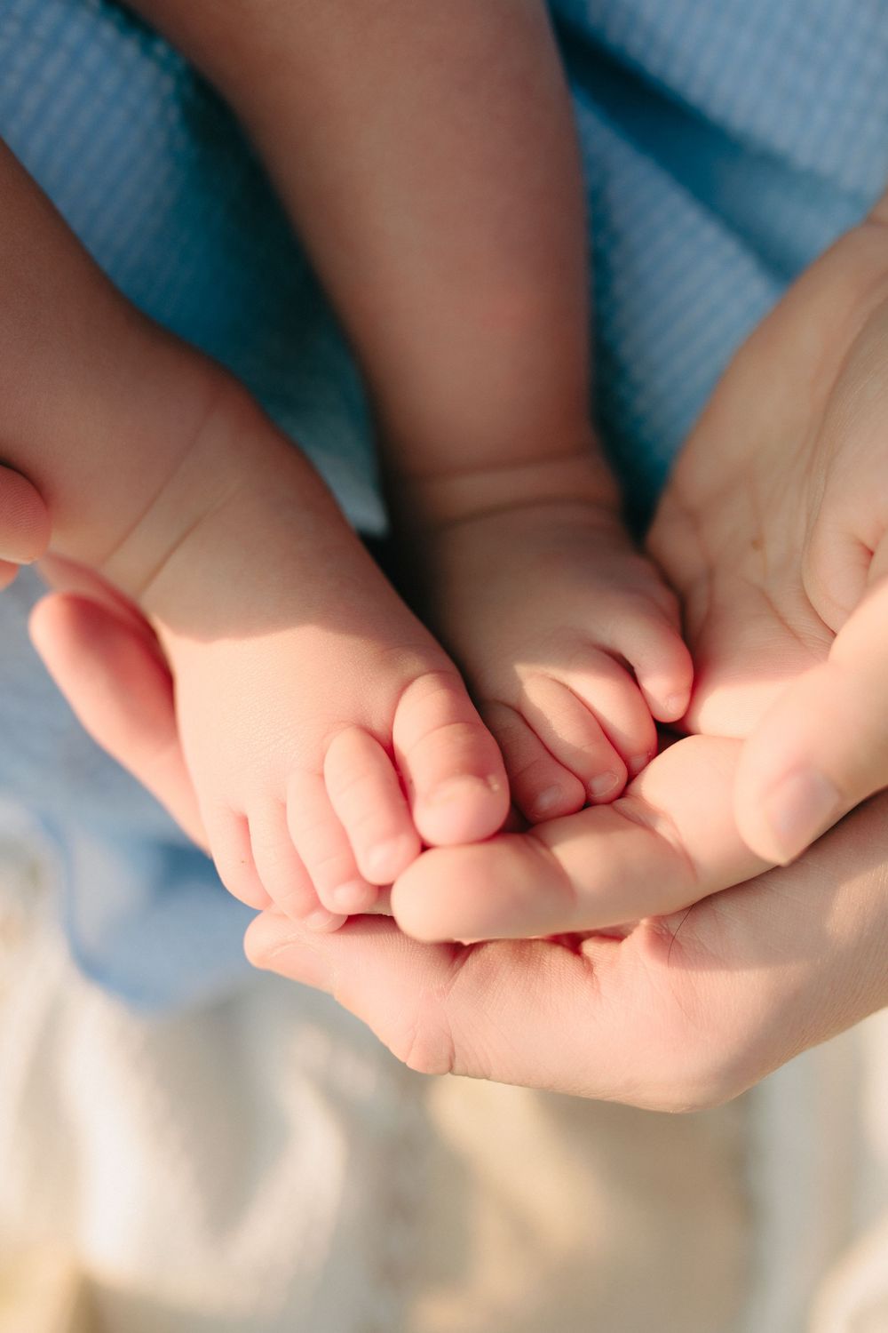 Adult hands gently cradle tiny baby feet against soft blue fabric background.
