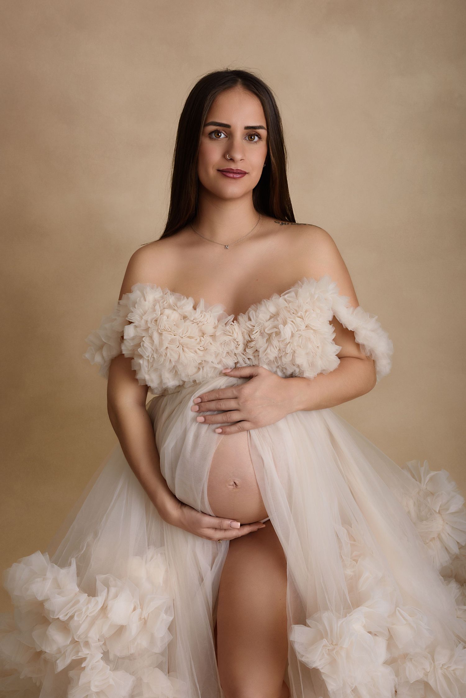Maternity photoshoot featuring an elegantoff-shoulder gown with tulle ruffle details against a neutral backdrop.