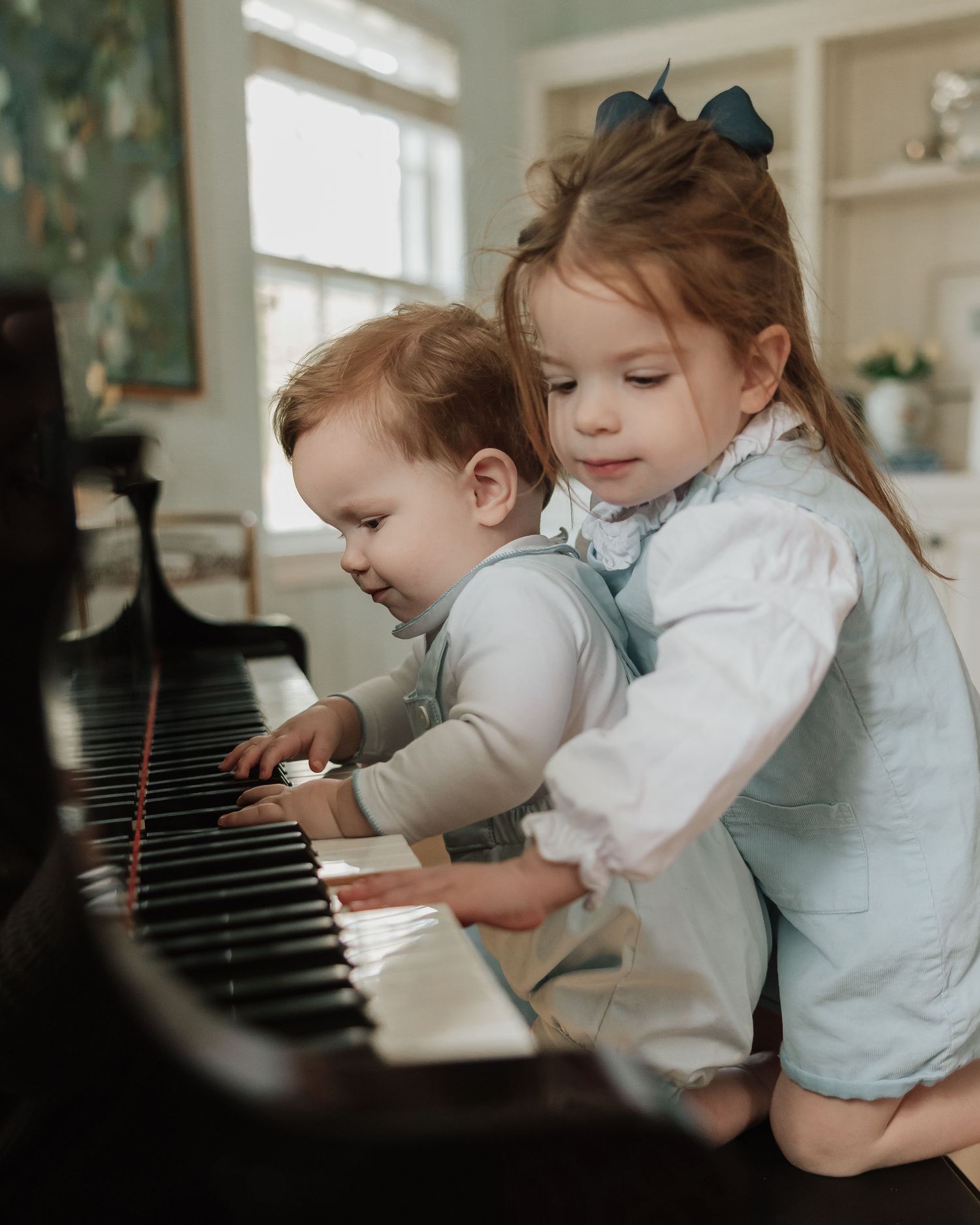 Two children sit together at a piano, with the smaller one pressing keys while the older one watches and helps.