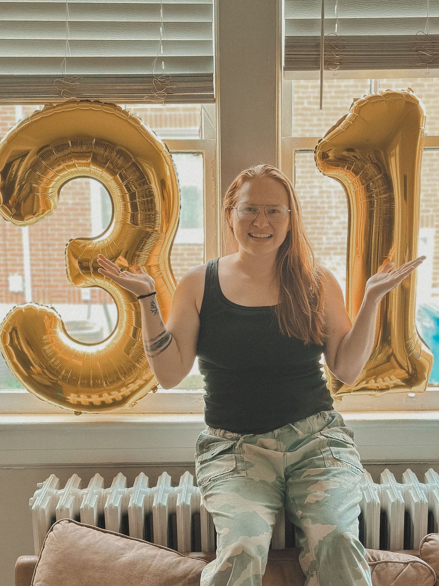 Woman smiling between gold "31" balloons, wearing camo pants and a black tank top, sitting on a radiator in front of a window.