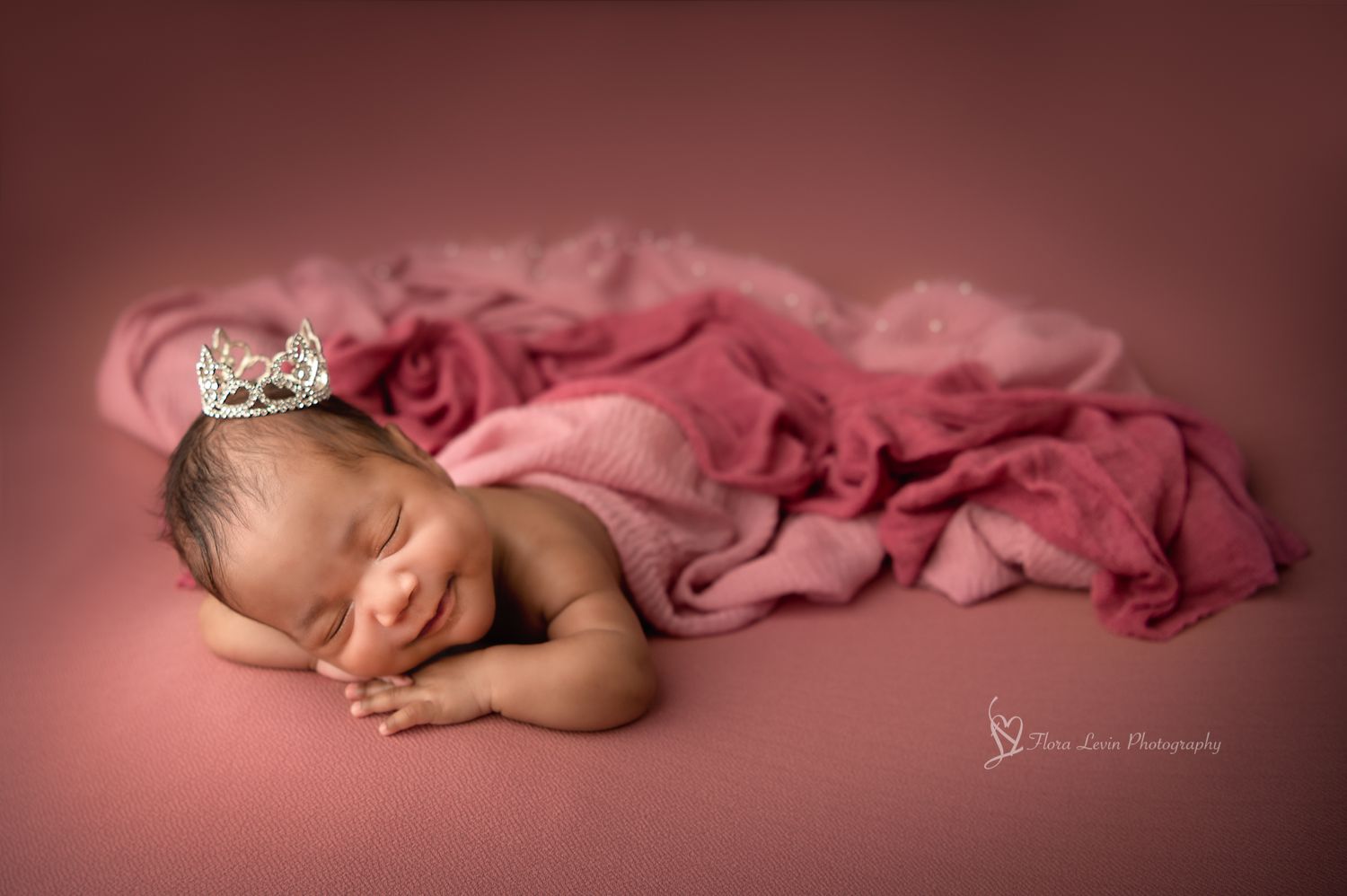 Sleeping newborn wrapped in soft pink fabric wearing a delicate crown in professional studio portrait.
