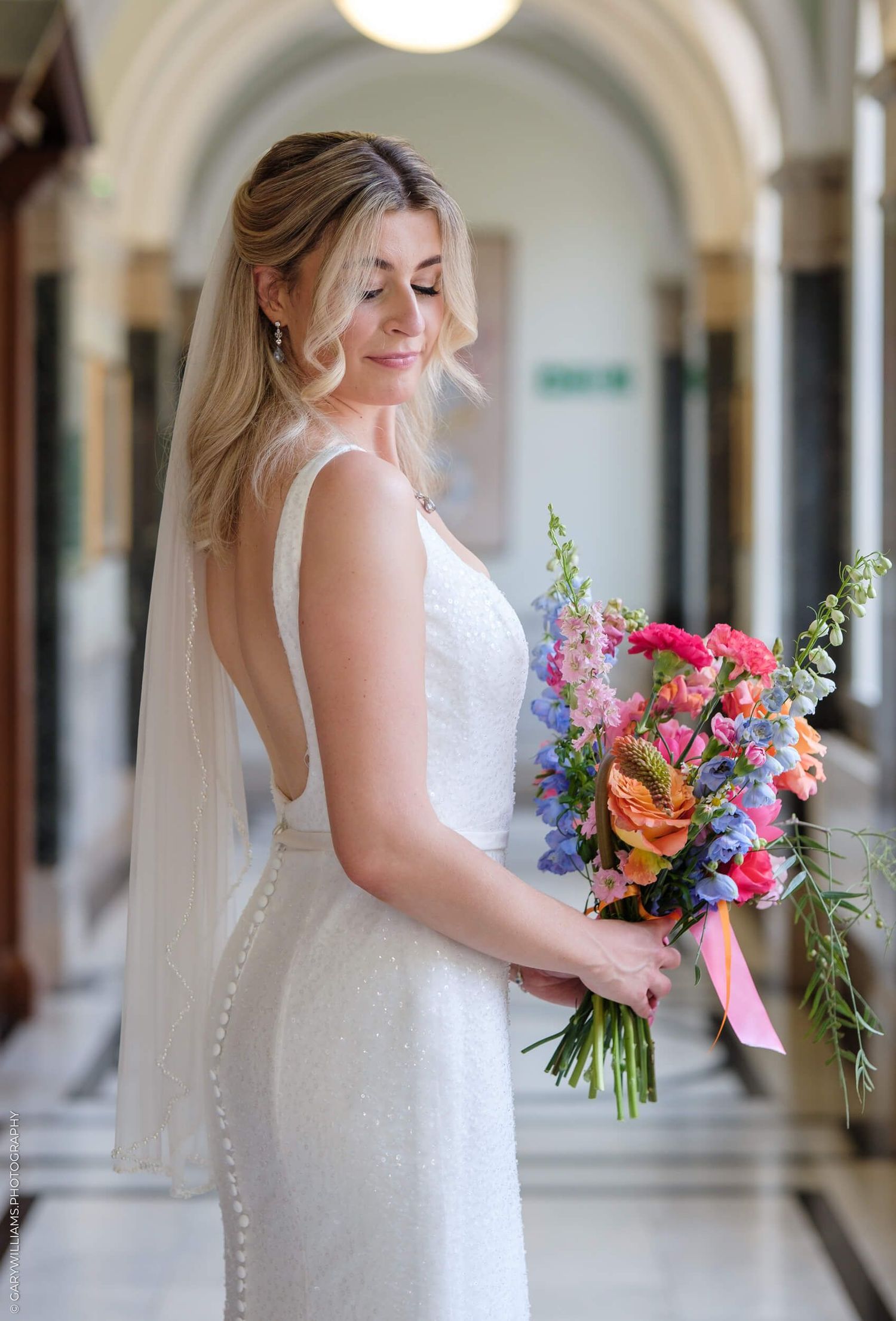 A bride in a white fitted dress holds a vibrant wildflower bouquet while standing in an elegant hallway corridor.
