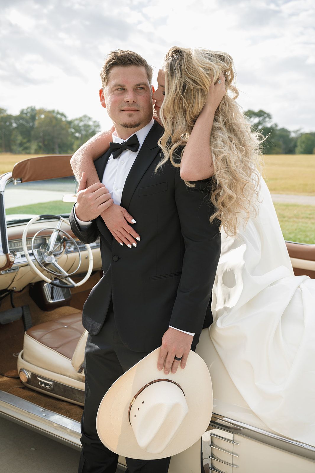 Bride and groom pose by vintage car in rustic outdoor setting with cowboy hat in hand.