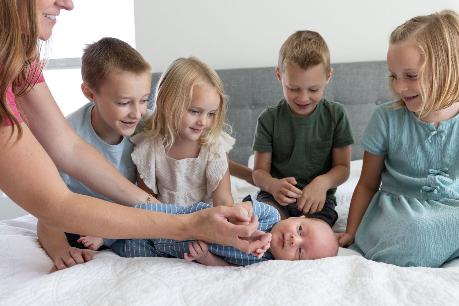 5 kids sitting on a bed looking at newborn baby with mom adjusting baby to face the camera