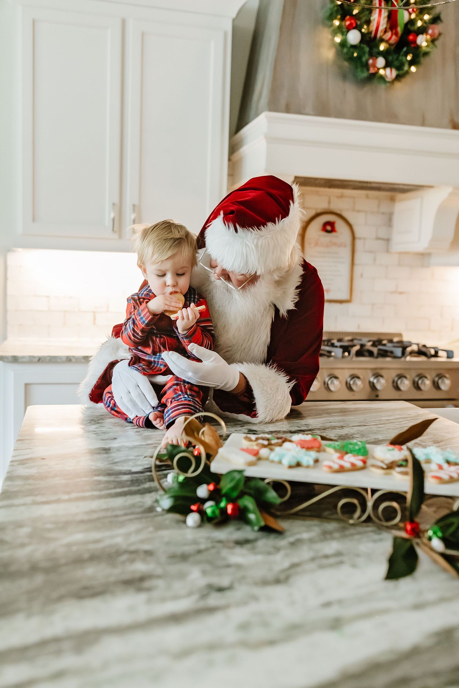 Holiday scene with cookie decorating on marble countertop with festive wreath on wall and Christmas decor.
