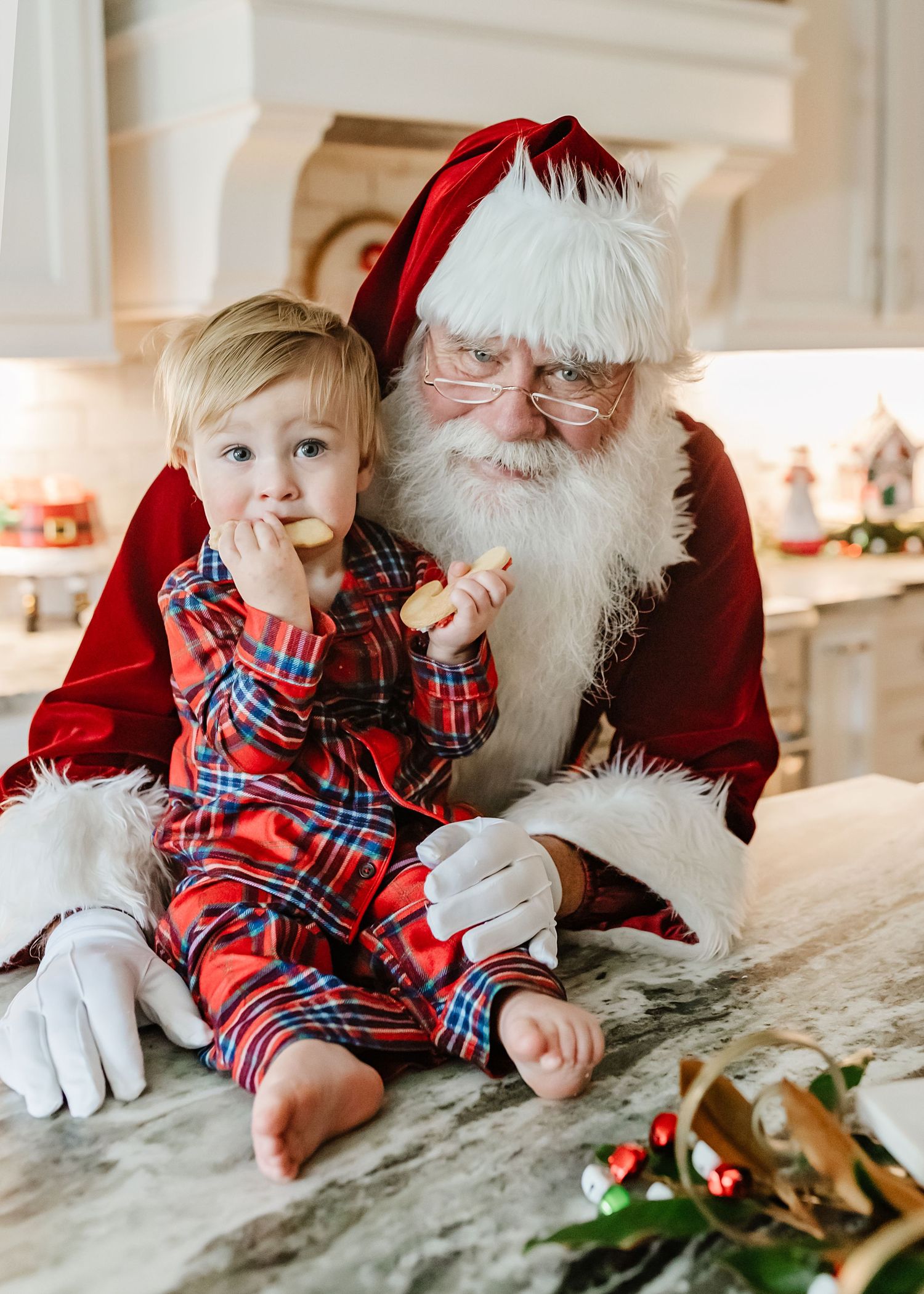 Baby in red plaid pajamas sits with Santa Claus during Christmas photoshoot on white bedding.