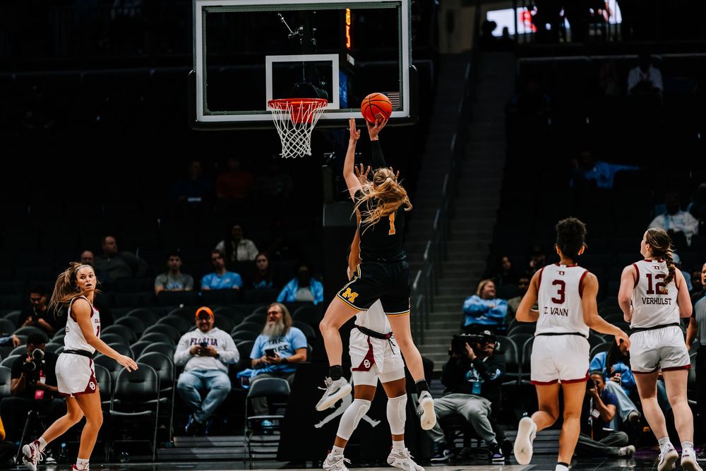 Women's college basketball player shoots layup while defenders watch during arena game.