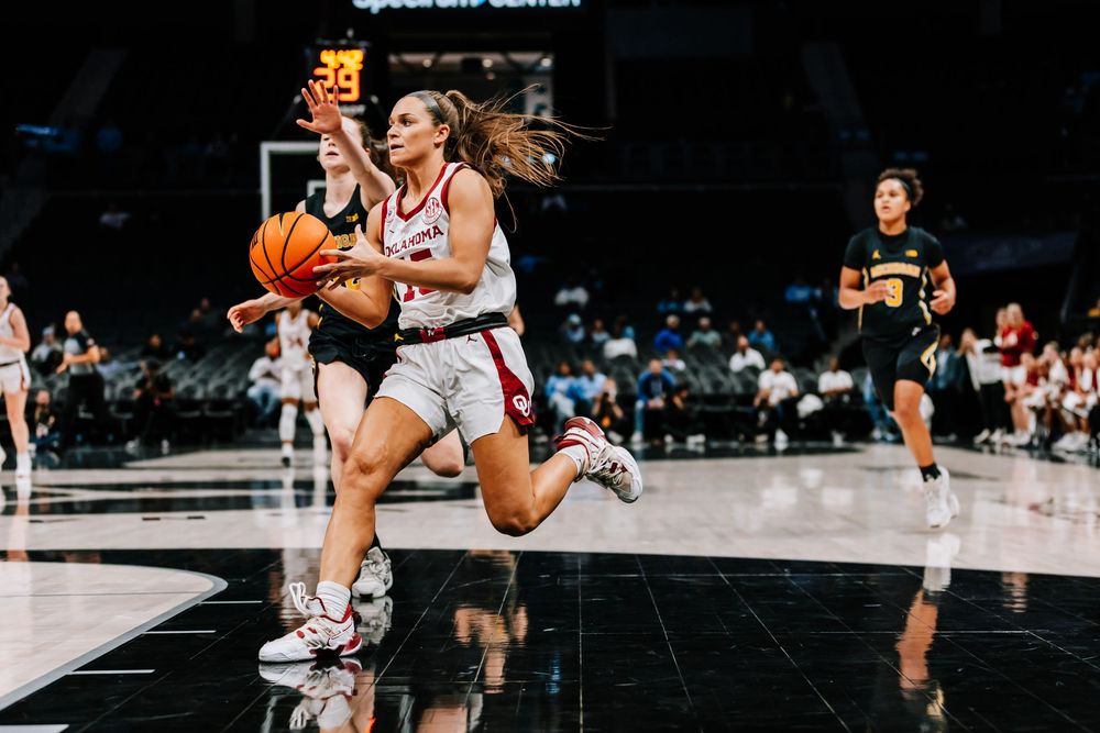 Female basketball player in white uniform drives to basket during women's college basketball game.