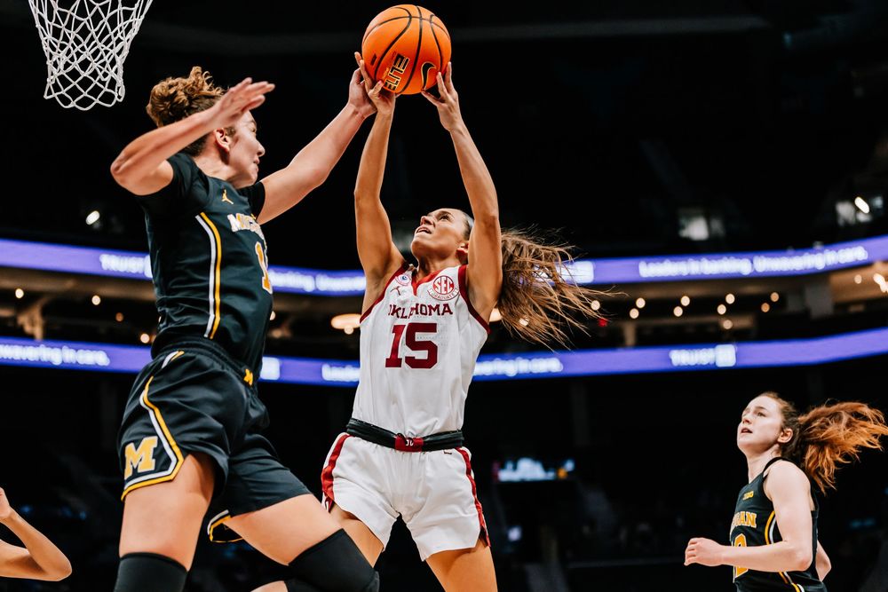 Oklahoma player #15 drives to the basket against a Michigan defender in a women's college basketball game.