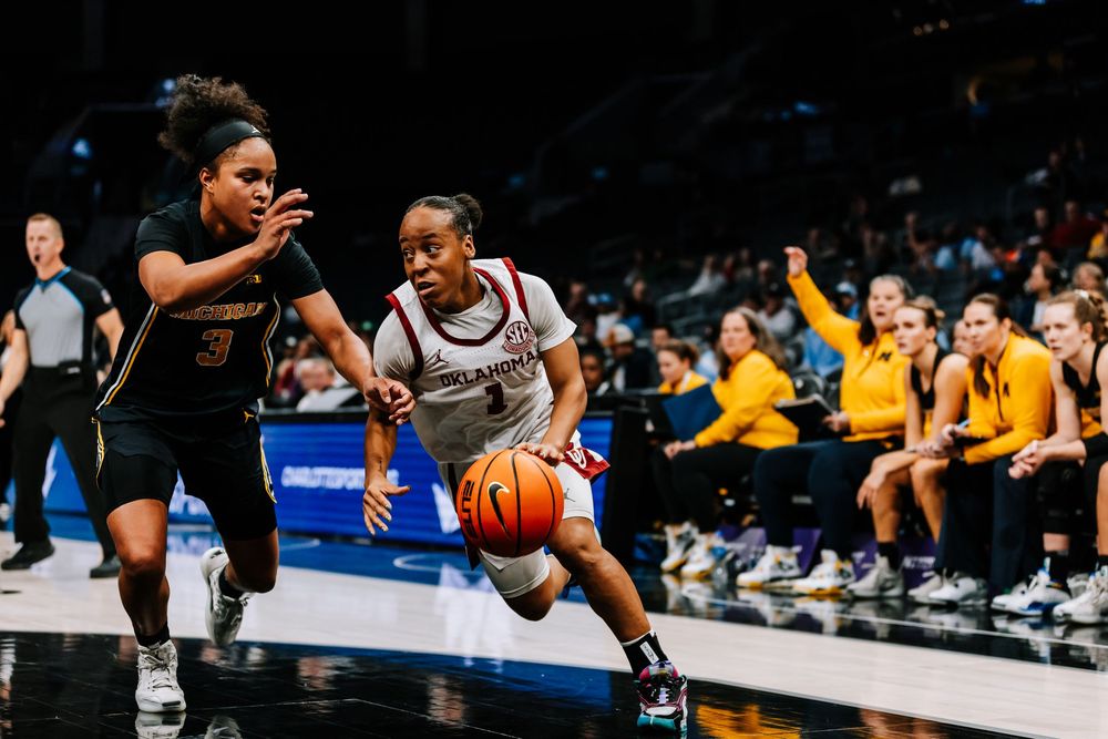 Oklahoma women's basketball player drives past a Michigan defender during a college game.