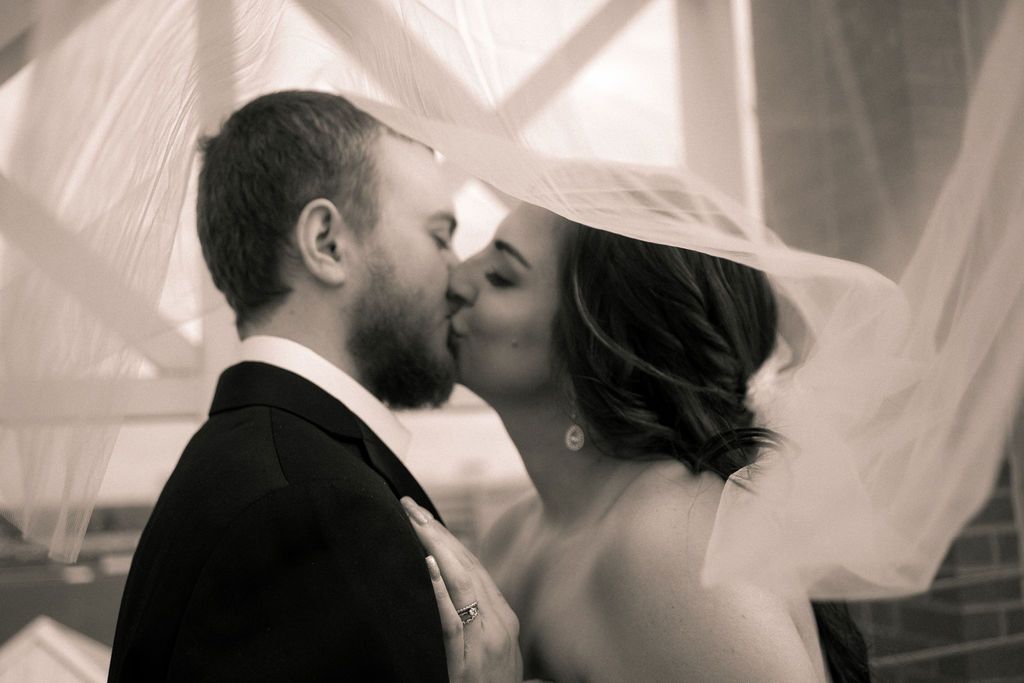 A romantic black and white photo of a couple sharing a kiss under a flowing veil during their wedding ceremony.