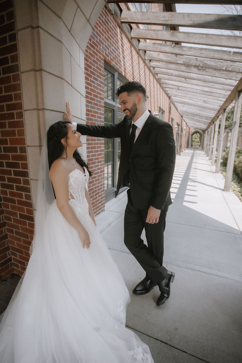 A wedding couple shares a tender moment in a sunlit covered walkway with brick walls.