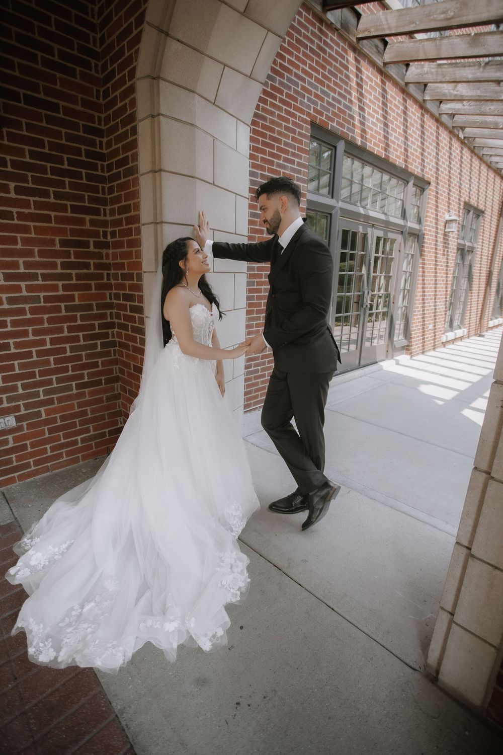 A couple shares a playful moment in formal wedding attire beneath an elegant brick archway.
