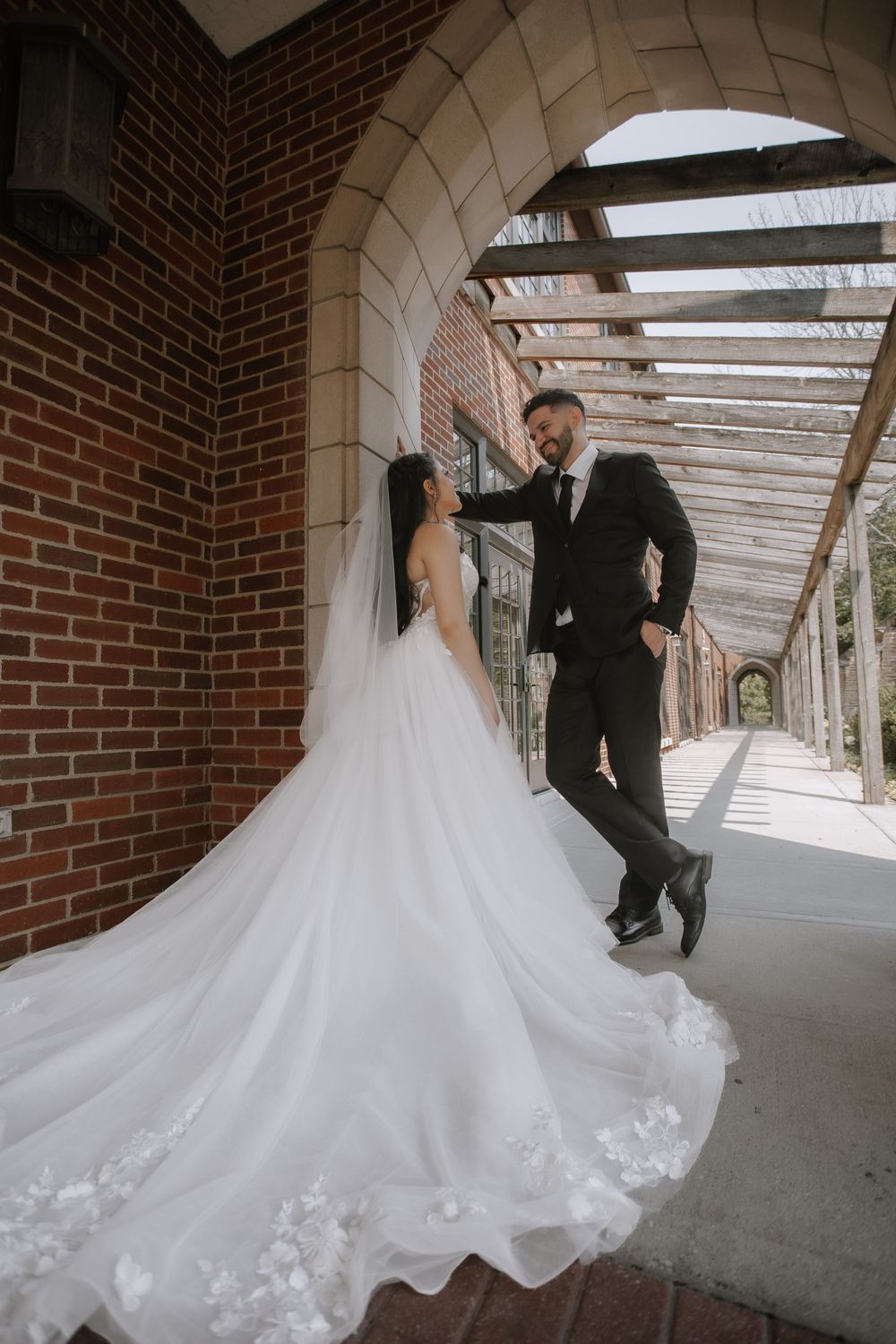 A bride in a flowing white gown twirls playfully in a covered brick archway walkway during a wedding photo session.