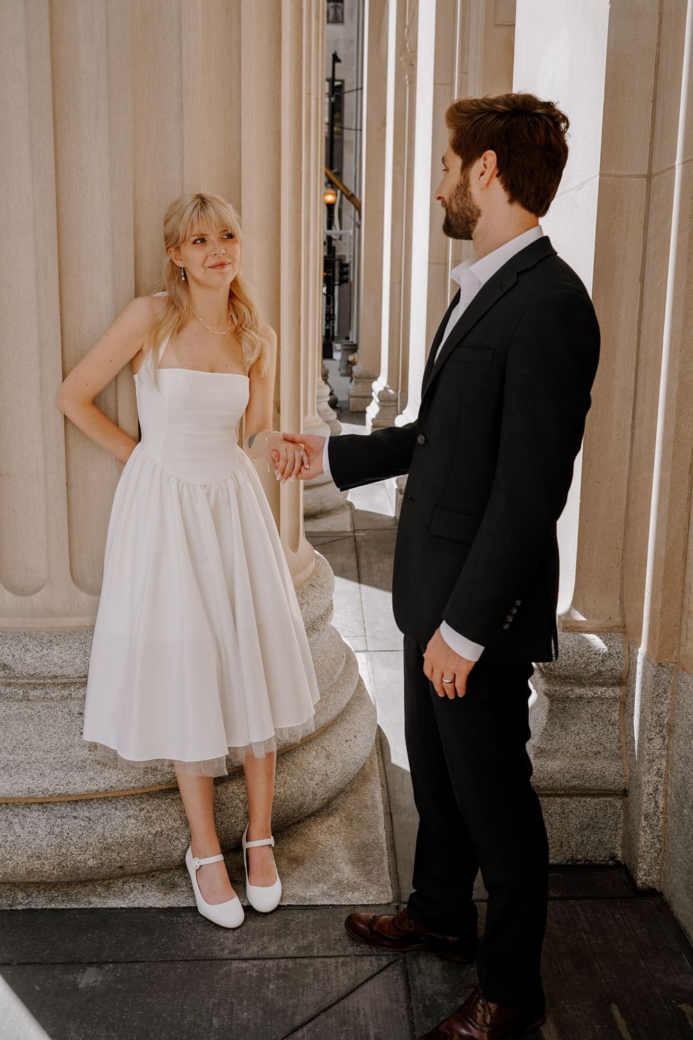 A couple in formal attire poses in an elegant columned corridor for artistic wedding photos.